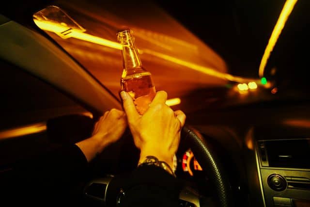 a close up of a drivers hands holding a bottle of beer and smoking a cigarette while holding onto the steering wheel of their car.