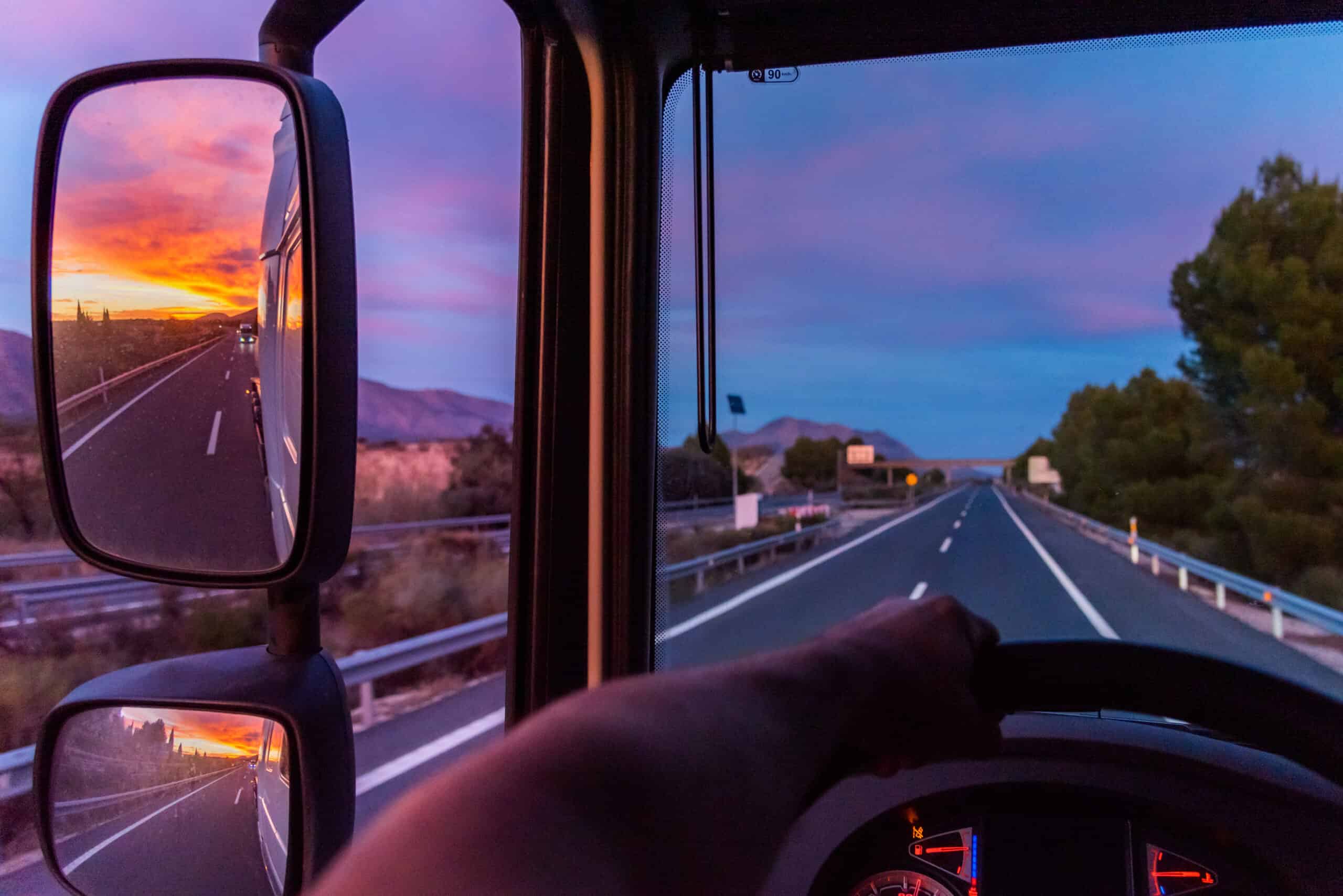 the sun sets in the rear view mirror of a commercial tractor trailer truck as it drives down the highway.