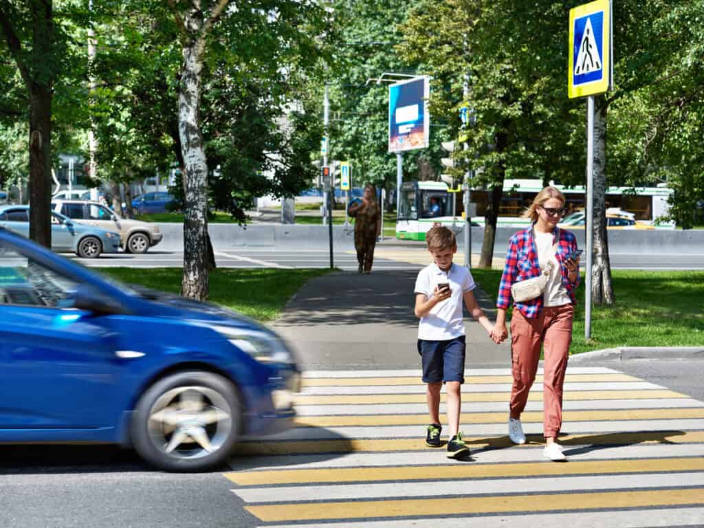 A woman and her son cross the road at a pedestrian crosswalk with a blue car approaching them from the side.