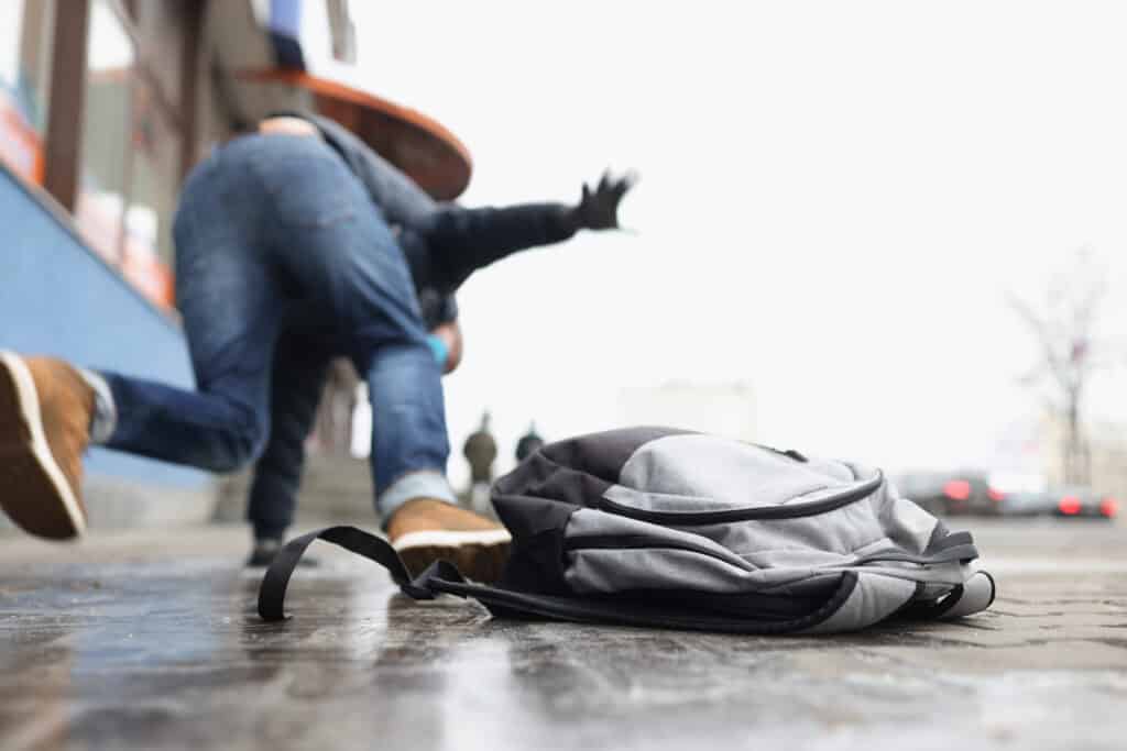 A person drops their backpack on the ground as they slip and fall on some ice on the sidewalk. 